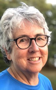 A woman, Eileen Harrington with short grey hair and dark-framed glasses smiling brightly for a headshot picture outdoors wearing a blue t-shirt.
