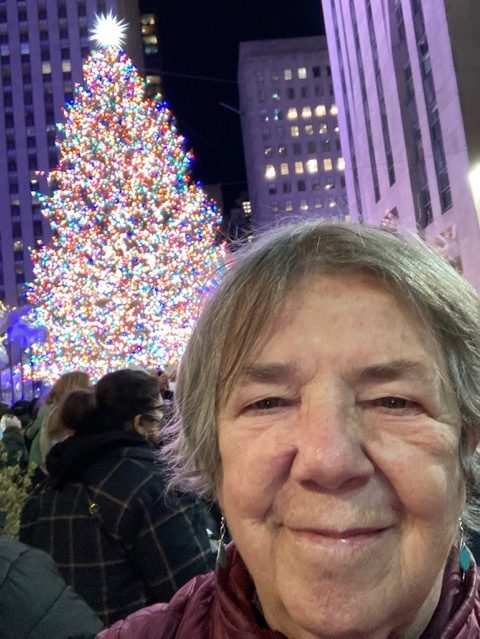 A woman takes a selfie with a huge, beautifully lit Christmas tree in the square behind her.