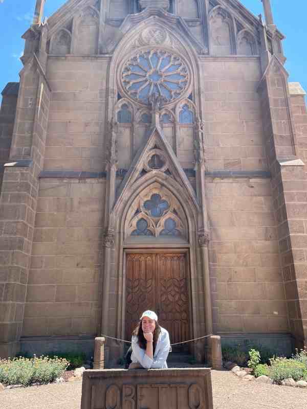 A young woman in a white baseball cap stands in front of a tall stone chapel with a rose window above the arch of the door. She leans on a carved stone sign that reads "Loretto."