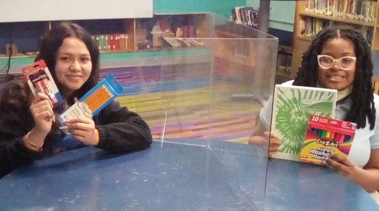 Two school girls smiling and displaying new school supplies in their classroom.