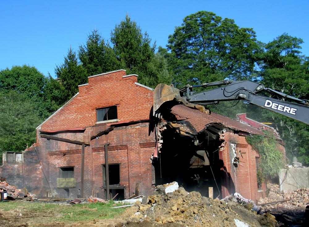 A red brick building being demolished by a large Deere excavator outdoors in front of large green trees and a blue sky.