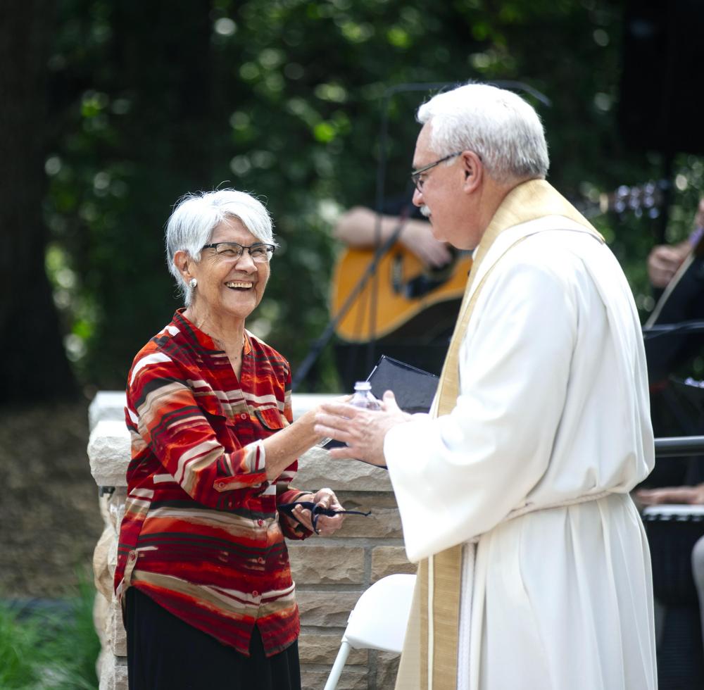 A pastor, Father John Fitzgibbons, and a woman, Sister Lydia Peña, greeting one another happily at an outdoor event with a live band behind them.