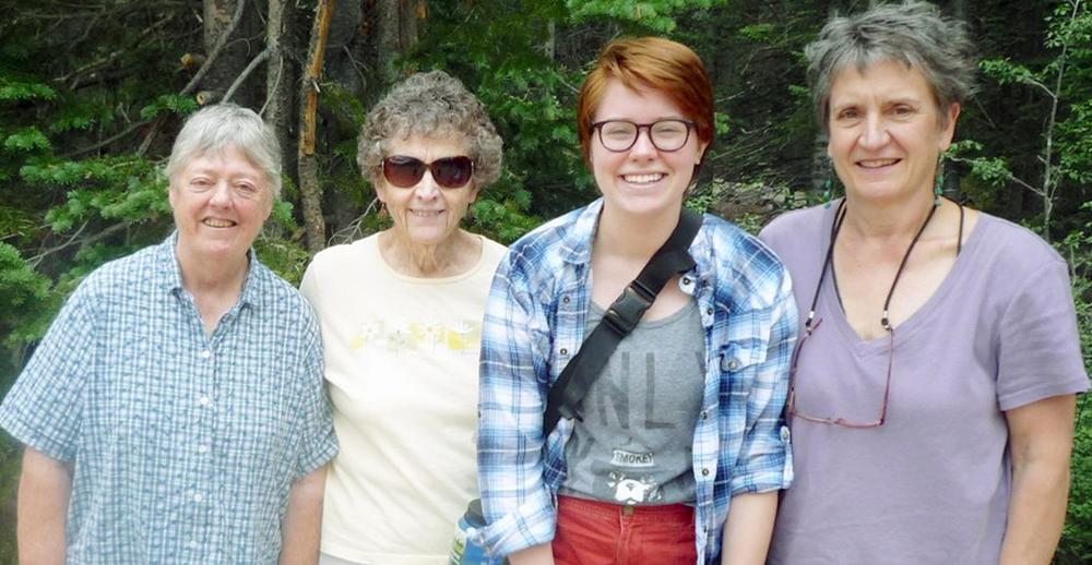 Four women stand together and smile for a group picture outdoors with thick green trees and brush behind them.