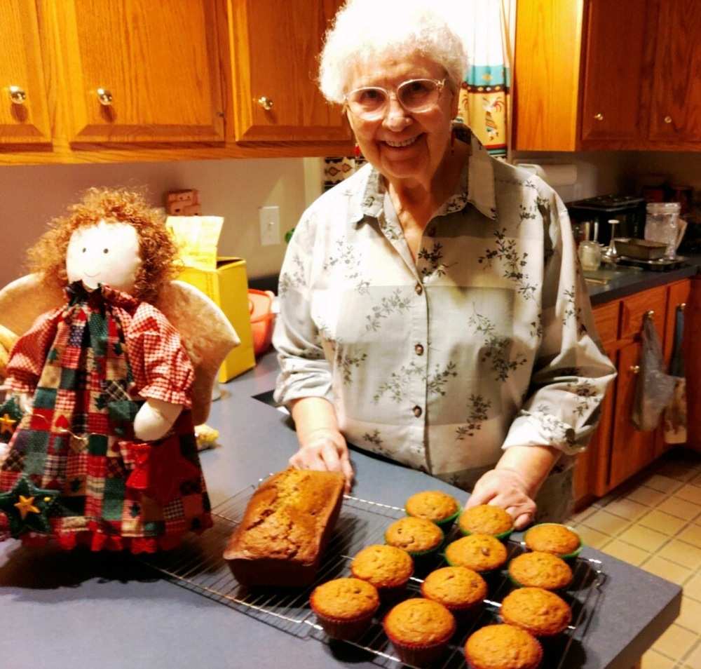 A smiling woman stands at her kitchen counter with a rustic angel doll next to cooling rack with a loaf of bread and muffins.