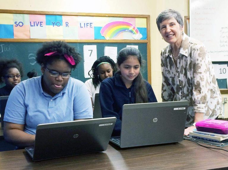 Classroom of young girls using computers with a teacher smiling at the camera.