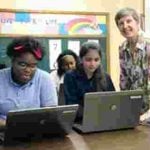 Classroom of young girls using computers to learn with a teacher smiling at the camera.