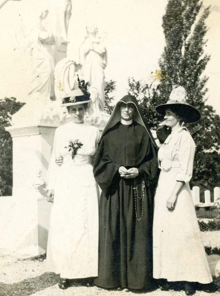 Archival photo of a nun standing between two women in long dresses and hats. A statue is in the background.