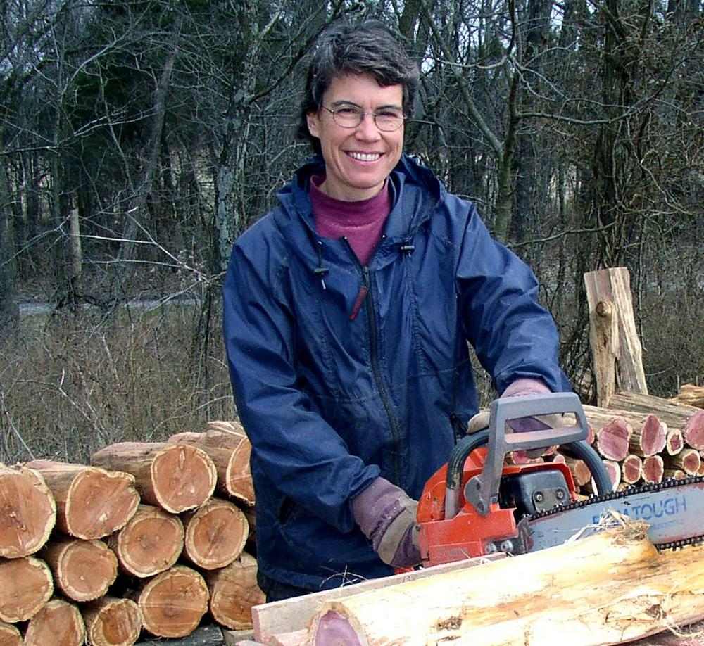 Woman holding a chainsaw partway through a log with logs neatly corded behind her.