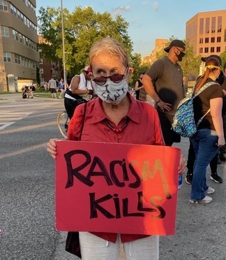 Woman in sunglasses and mask stands in the street holding a red sign that reads "Racism Kills."