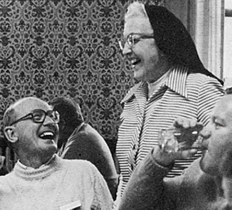 Archival black and white photo of a nun in a veil standing next to two men who are seated. One is drinking from glass; the other is looking up at the nun as they share a laugh.