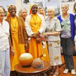 A diverse group of religious women, from Ghana and the US standing and smiling together for a photo at a committee meeting.