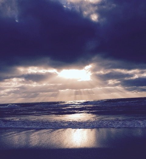 The sun streams down through an opening in the clouds to illuminate the waves striking the beach.