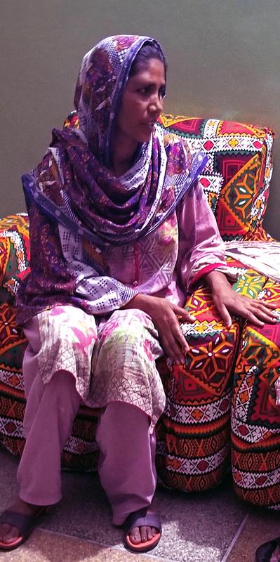 A woman in a colorful headscarf and traditional Pakistani dress sits on the couch talking with someone out of the camera shot.