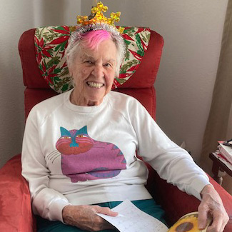 A woman with short white hair with a bright pink stripe in the center wearing a crown headband and a white sweater with a colorful cat graphic smiling brightly for a picture while sitting in a red recliner chair indoors and holding papers in her hands.