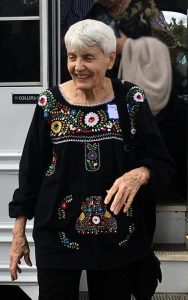 A woman in a black top dress embroidered with colorful flowers steps off a white bus.