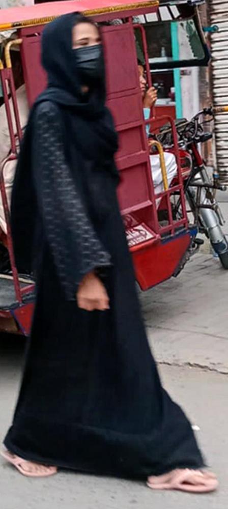 A woman dressed in black, with a black scarf around her head and a black mask, walks down the street.