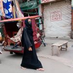 A woman dressed and wearing a black headscarf and mask walks down the street in Pakistan.