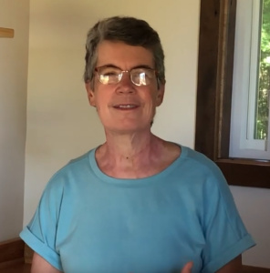 A woman standing near a window inside a simple room, smiles.