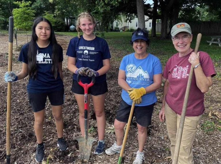 Two high school girls holding garden tools stand outside in mulch next to two women, also holding garden tools, for a photo.