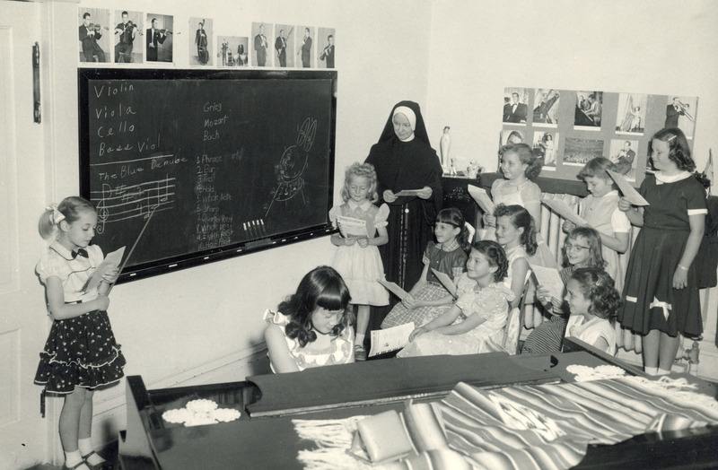 A nun in a habit watches over her class of young girls who appear to be singing from sheet music while one girls accompanies on piano.