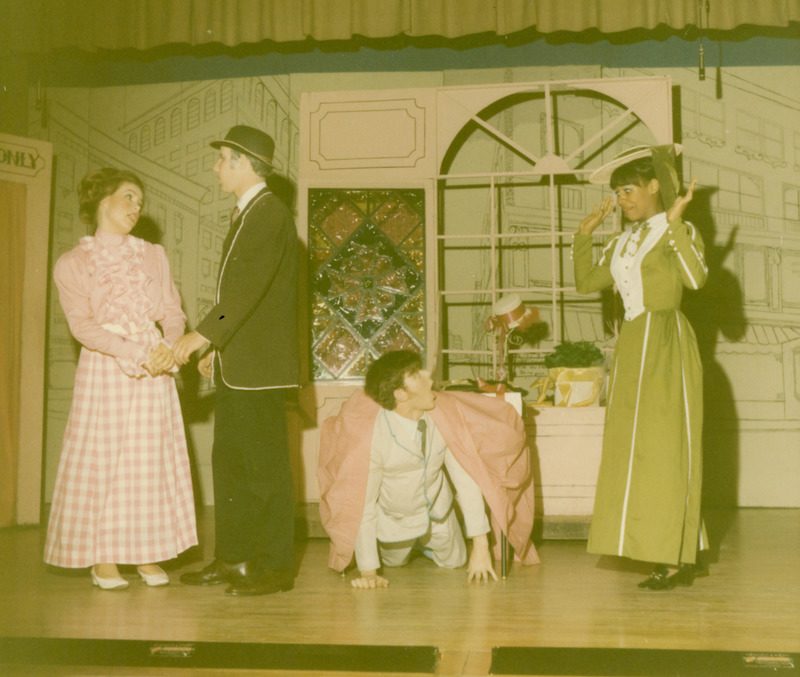 A moment in a play - a young couple in love are holding hands and singing to each other, and a young man coming out from under a table looks up in shock at a surprised young woman who has discovered him.