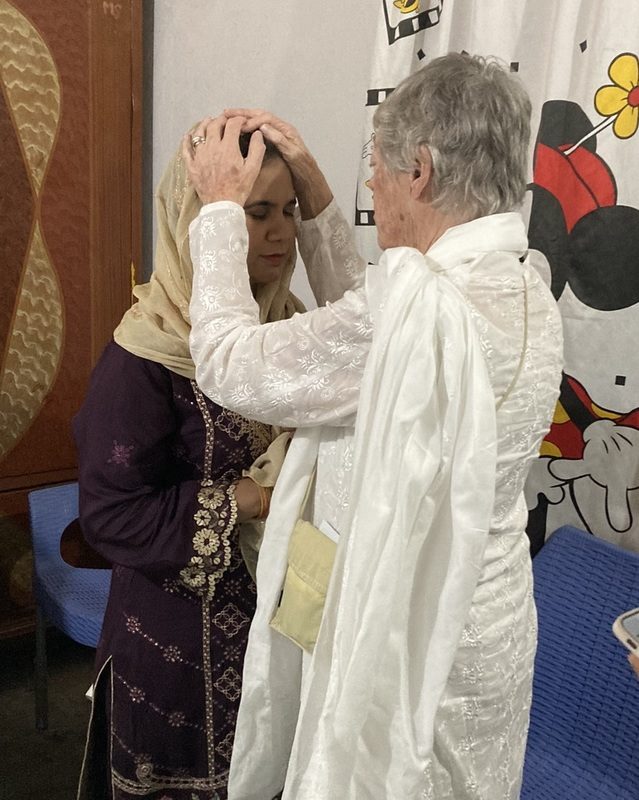 A woman in a white tunic places her hands in blessing on the head of a woman wearing a headscarf and embroidered tunic.