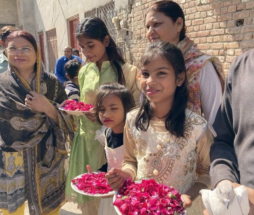 Three young girls in fancy dresses hold plates of rose petals in Pakistan. Another person holds a pigeon to give to the guests of honor to release.