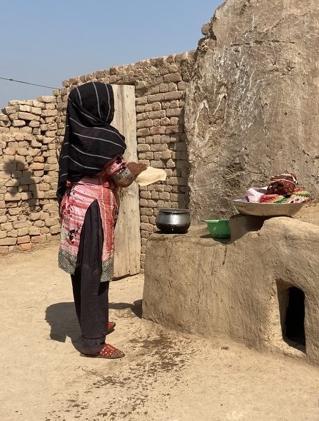 A woman in a tunic and headscarf holds a thin bread in her hand at an outdoor kitchen.