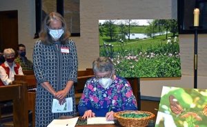A woman sitting down signing paperwork as another women watches over. They're surrounded by beautiful scenery photography and individuals sitting behind them.