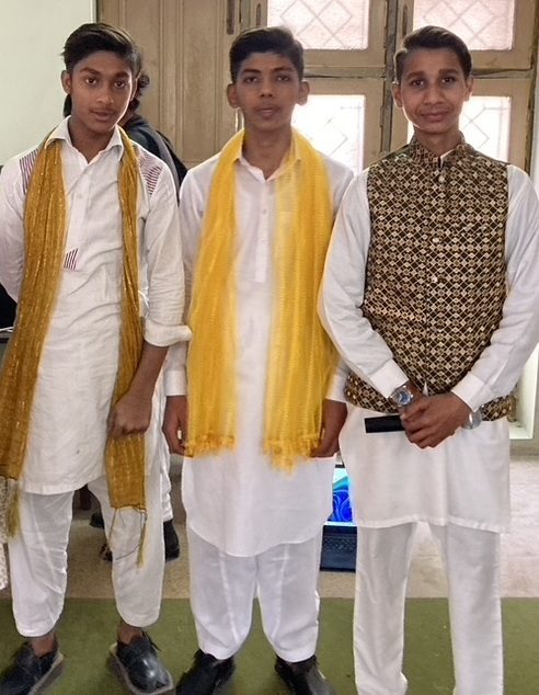 Three young men in white tunics and trousers pose for a photo. Two of them wear long gold/yellow scarves, and one sports a patterned vest.