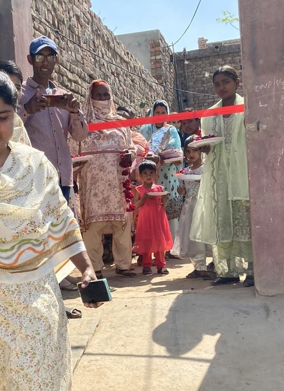 A group of people of all ages wearing Pakistani tunics and holding plates of rose petals wait at an entrance.