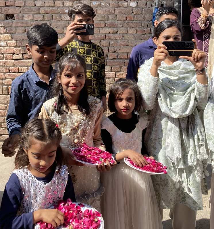 Young Pakistani girls in fancy dresses stand with plates of rose petals, while older teens hold cells phones to document the moment.