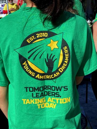 A girl standing outside with her back facing the camera to show the back of the green t-shirt she is wearing. The t-shit says, "Young American Dreamers: Tomorrow's Leaders, Taking Action Today.