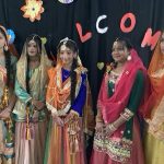 Five young women in vibrantly colored traditional Pakistani dress pose for a photo. They all wear colorful headscarves and their clothes are embroidered with gold thread.