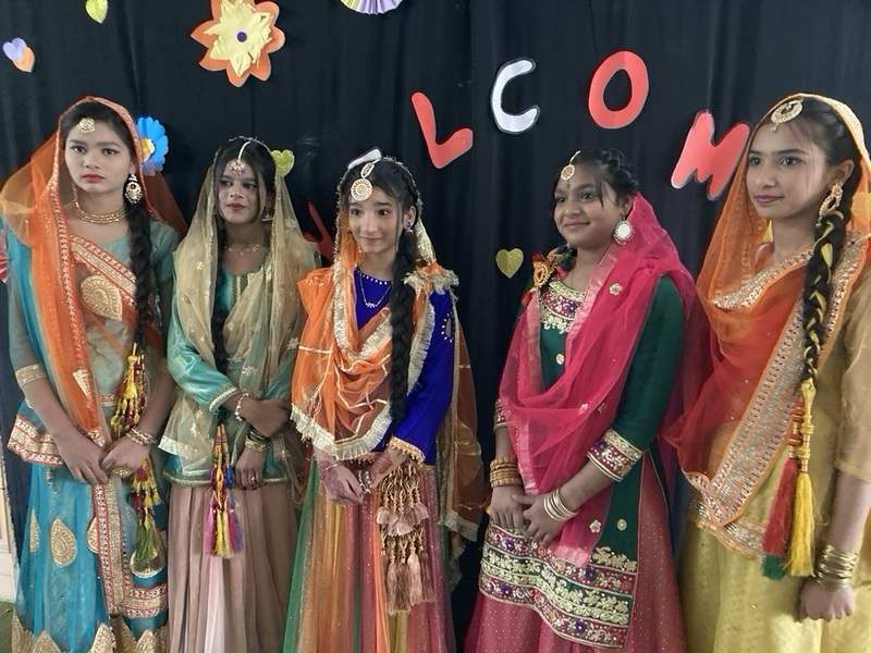 Five young women in vibrantly colored traditional Pakistani dress pose for a photo. They all wear colorful headscarves and their clothes are embroidered with gold thread.