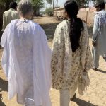 Two women and two men in Pakistani tunics walk down a dusty brick road.