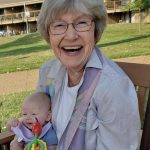A woman with a beaming smile holding a young infant on her lap sits in the green space of an apartment complex.