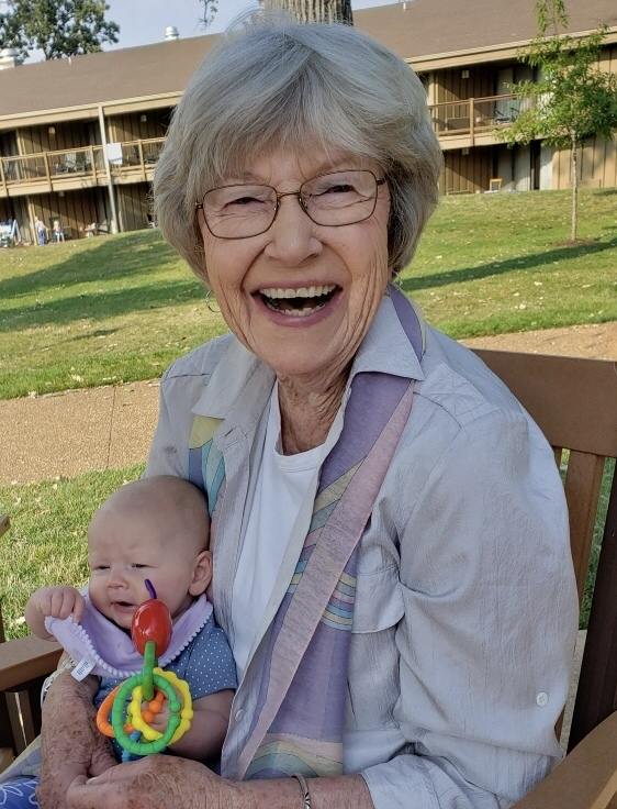 A woman with a beaming smile holding a young infant on her lap sits in the green space of an apartment complex.