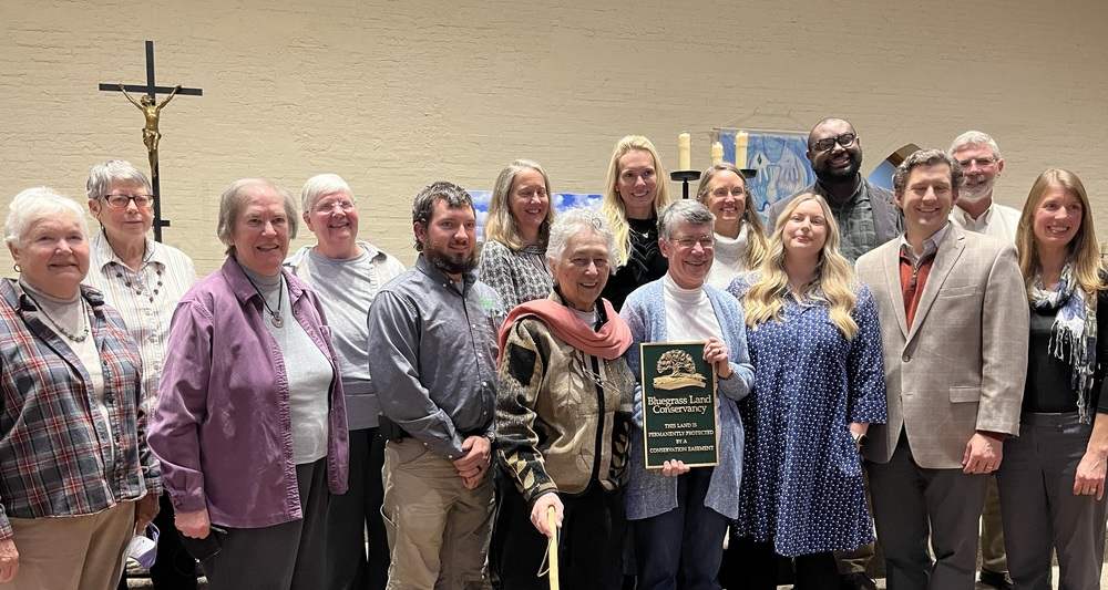 A large group of people pose for a group photo, with a crucifix visible in the background and the center person holding a plaque with the header "Bluegrass Land Conservancy."