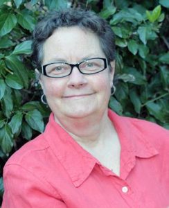 Short-haired woman in pink collared shirt smiling for a head shot in front of greenery.