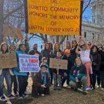 A large group of individuals collected outdoors for a photo with a sign saying, "Loretto Community honors the memory of Martin Luther King Jr.
