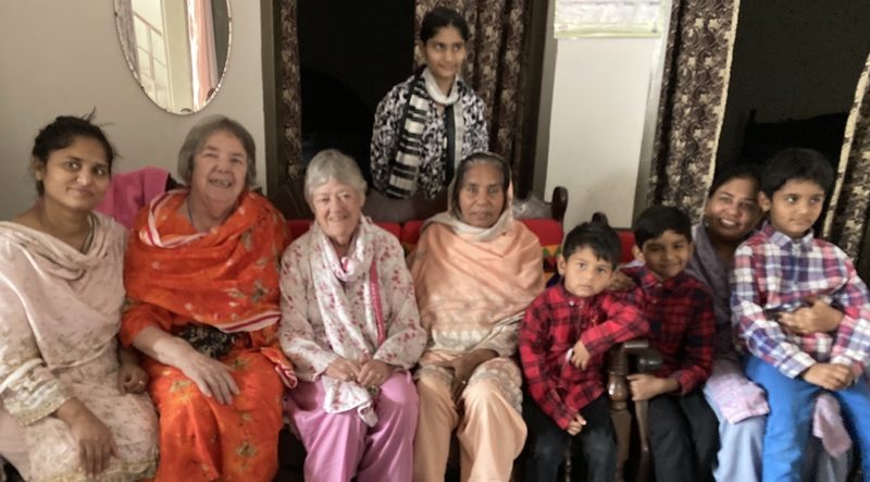 A group of nine people, some wearing shalwar kameezes, sit together and smile for a photo. The woman on the right side of the photo has a little boy sitting on her lap, and two others on her left.