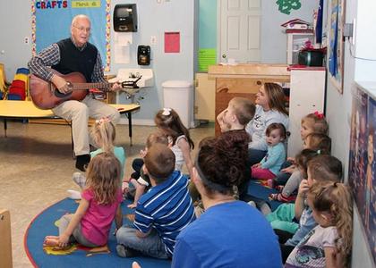 A man in a plaid shirt and sweater vest playing a guitar for a classroom of children and two teachers all sitting on the ground.