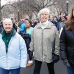 Three women smiling together in cold weather clothing walking in a crowd down a street for the womens march.