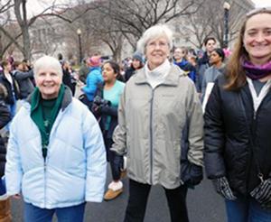 Three women smiling together in cold weather clothing walking in a crowd down a street for the womens march.