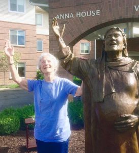 A short haired woman in blue shirt stands next to a brass statue and mimics the pose on a sunny day.