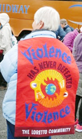 Short haired woman displaying a handmade colorful red cape that says: Violence has never defeated violence.
