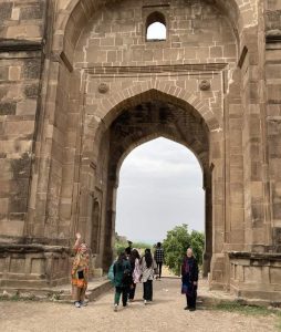 A woman stands, waving, at the gate in an old stone wall as other people walk through it.