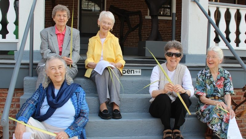 Five women sit on the steps of a porch, holding palms and smiling.