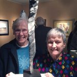 Two women smiling with a small silver sculpture at a gallery event inside.